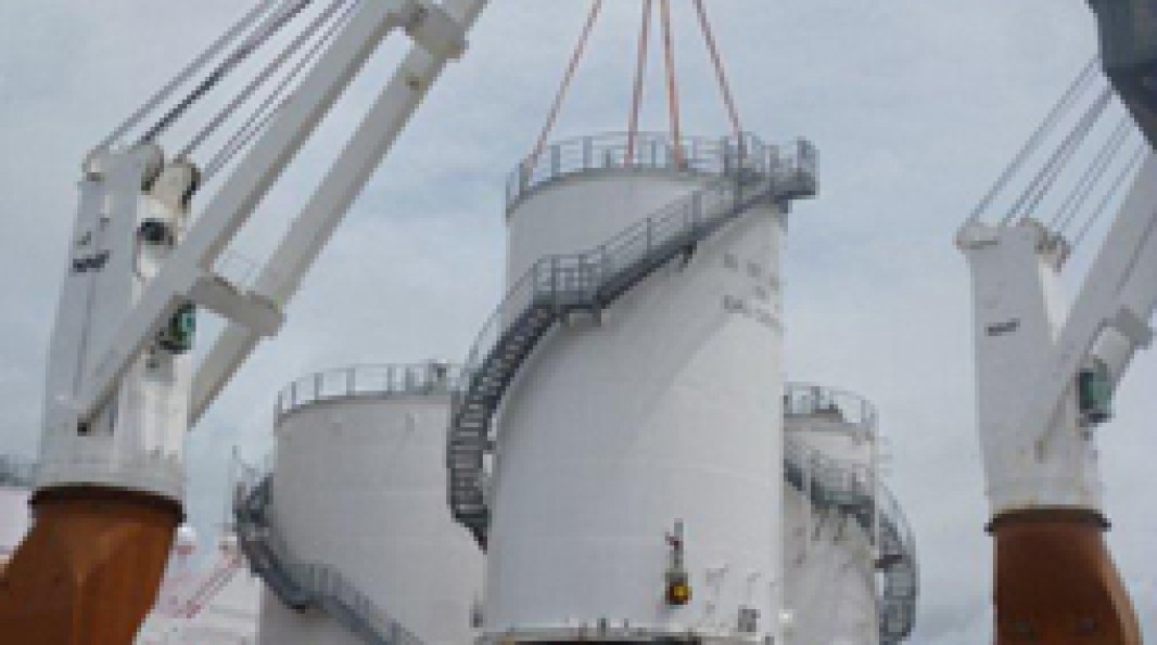 Photo du levage d’une cuve cylindrique blanche à l’aide de câbles, entourée par des bras de grues portuaires