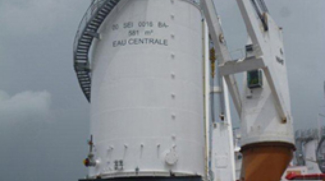 Photo zoomée d'une cuve marquée "EAU CENTRALE", suspendue en l'air par une grue, lors de son transfert vers le site de la centrale