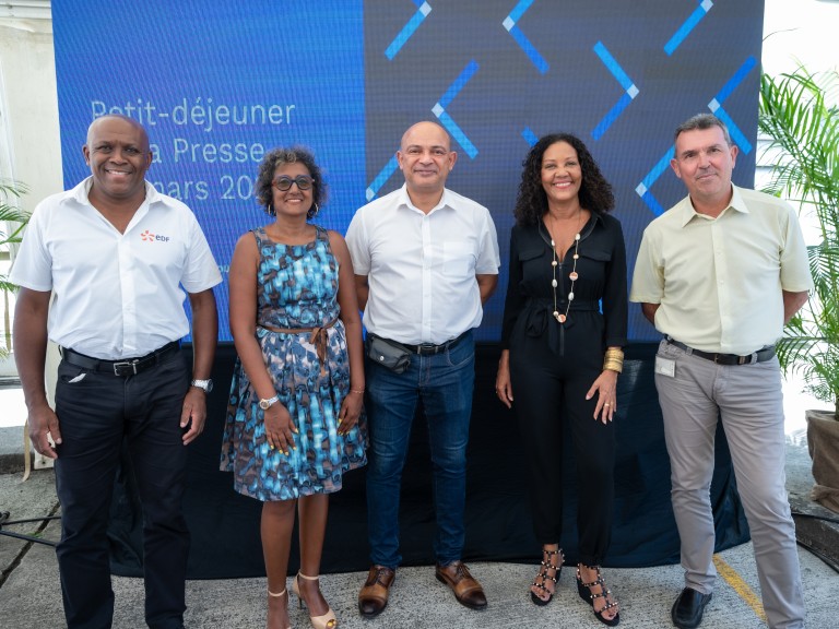 Photo de cinq représentants d’EDF Archipel Guadeloupe posent devant un fond bleu affichant 'Petit-déjeuner de presse - mars 2024'